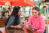 Optimistische junge Freundinnen in legerer Kleidung nutzen Smartphones und diskutieren über Nachrichten, während sie an einem sonnigen Tag auf der Veranda eines Restaurants sitzen