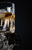 Whiskey with ice cubes served in crystal glass placed on rough surface against black background
