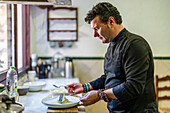 Seitenansicht eines konzentrierten männlichen Kochs in schwarzer Uniform mit Armbändern, der in einer modernen Restaurantküche schmackhafte Speisen auf einem Teller serviert
