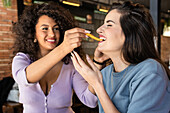 Positive junge ethnische Frau mit dunklen lockigen Haaren füttert hungrige fröhliche Freundin mit appetitlichen Pommes frites im Restaurant