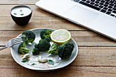 Leckerer gekochter Brokkoli mit Zitronenscheibe und Cashewnüssen neben Schüssel mit weißer Soße und Netbook auf Holztisch