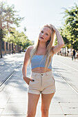 Charming female with blond hair and in trendy summer clothes standing with hand in pocket in city and looking at camera