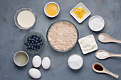 Top view of different ingredients including milk melted butter near bowls of lemon zest flour sugar salt near sour cream soda oil and blueberries eggs with flavoring for pastry recipe
