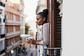 Seitenansicht einer nachdenklichen Afroamerikanerin mit einer Tasse Heißgetränk, die von einem Balkon in der Stadt wegschaut