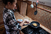 From above side view of focused young Asian female in casual clothes cooking yummy pancakes using spatula while standing in kitchen