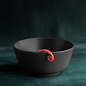 Curled octopus leg coiling out of grey bowl on plain dark background.
