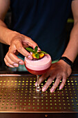 Hände eines nicht erkennbaren Barkeepers, der einen Cocktail mit Minzblättern und Passionsfrucht in der Bar dekoriert