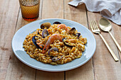 Teller mit leckerer frischer Paella mit Muscheln und Krabben, serviert auf einem Holztisch mit einem Glas eines erfrischenden Getränks und Besteck