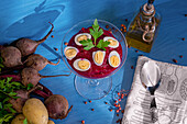 From above glass goblet of tasty beetroot cream garnished with quail eggs and parsley placed near napkin with spoon and fresh ingredients against blue background