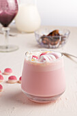 Glass of sweet hot white chocolate with pink jelly candies and marshmallow served on white table