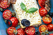 Close up of ripe cherry tomatoes with black olive slices and feta cheese in baking dish near uncooked penne