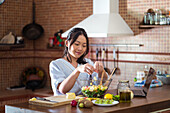 Delighted young Asian woman with long dark hair in casual clothes smiling while standing at table with laptop and ingredients and preparing delicious salad in kitchen