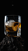 Whiskey drops falling on ice cubes served in crystal glass placed on rough surface against black background