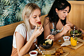 Young content best female friends sitting at wooden table with tasty croissants and beverages in cafeteria