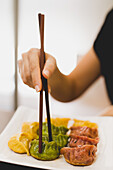 Hand of woman holding bamboo chopsticks and eating colorful dumplings from white ceramic square plate