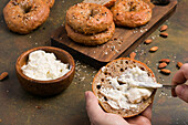 Crop anonymous male spreading tasty cream cheese on low carbohydrate bagel with sesame seeds served on table with almonds and cutting board