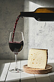 Semi sweet red wine pouring into wineglass placed on wooden table near triangle cheese piece in studio