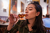 Attraktive Asiatin mit schwarzen Haaren trinkt während des Abendessens ein alkoholisches Getränk aus einem Weinglas, während sie in einem Restaurant mit Menschen im Hintergrund sitzt