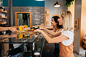 Side view of young cheerful best friends receiving trays with delicious food and beverages from crop anonymous vendor in cafeteria