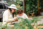 Mutter pflanzt Samen in Plastikschale auf Gartenbeet in Bauernhof mit liebenswertem Sohn