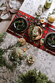 Top view of Christmas table setting with wreath on the plate, decorative wooden ornaments and red checkered tablecloth with yellow lights on the background