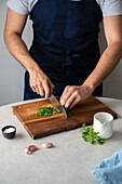 Unrecognizable male in apron cutting fresh parsley on wooden cutting board near salt and garlic while cooking lunch at home