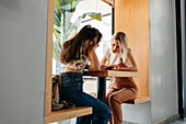 Anonymous best female friends surfing internet on cellphone while talking at cafeteria table in daylight