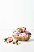 Leckere Eiskugeln auf einer knusprigen Waffelschale, dekoriert mit frischen, reifen Erdbeeren, Heidelbeeren, Pistazien und Minzblättern vor weißem Hintergrund