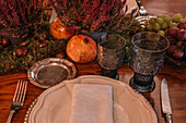 Kristallgläser in der Nähe von Teller und Besteck auf einem mit Weintrauben, Calluna vulgaris-Blüten und Granatäpfeln geschmückten Tisch