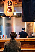 Back view of faceless black haired woman in sweater sitting at counter in cozy ramen bar