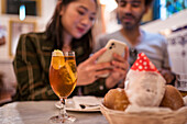 Crop young Asian female showing photos on smartphone to ethnic boyfriend sitting at table with glass of lemonade and bread basket in restaurant