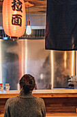 Back view of faceless black haired woman in sweater sitting at counter in cozy ramen bar