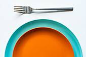 Von oben runde orangefarbene und blaue minimalistische Keramikteller, die neben einer silbernen Gabel auf einem weißen Tisch aufeinander gestellt sind