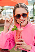 Verträumte Frau im rosa Pullover sitzt am Tisch mit einem köstlichen kalten Beerencocktail in einer Bar im Freien und schaut in die Kamera