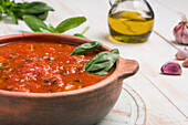 Holzschüssel mit roter Marinara-Soße aus Tomaten und Basilikumblättern auf dem Tisch, dazu Olivenöl und Knoblauch