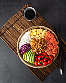 Top view of appetizing colorful poke bowl with fresh salmon and rice served with ripe cherry tomatoes avocado slices onion and mango placed on bamboo tray with soy sauce
