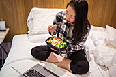 From above Asian female eating healthy salad and watching video on netbook while sitting on bed in light hotel room during quarantine