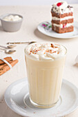 Glas Milchpunsch mit Zimtpulver auf geschlagenem Eiweiß vor Kuchenstück auf Cafeteria-Tisch vor hellem Hintergrund