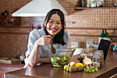 Hungry young Asian female with long dark hair in casual clothes eating appetizing healthy salad while leaning on counter in kitchen