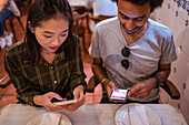 Happy young multiracial couple in casual clothes using smartphones while sitting together at table in modern restaurant