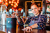 Fröhliche Barkeeperin mit einem Glas erfrischendem Wermut, die am Tresen einer Bar steht und in die Kamera schaut