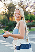 Side view of cheerful female standing with cold lemonade in plastic cup in street in summer looking at camera