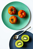 Draufsicht auf frische, gesunde halbierte Kiwi und ganze Tomaten auf blauen und türkisfarbenen runden Tellern mit Löffel auf weißem Hintergrund