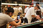 Waitress in uniform and protective mask taking photo on smartphone of chef and cooks preparing food in open kitchen during coronavirus outbreak