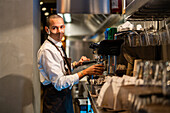 Seitenansicht eines Kellners in Uniform und Schutzmaske, der die Knöpfe einer Kaffeemaschine drückt, während er ein Getränk in einem Restaurant während einer Coronavirus-Pandemie aufbrüht