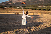 Female throwing hat and jumping along dried field in summer evening in countryside