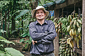Confident Hispanic man in shirt and bucket hat crossing arms and looking at camera while standing near shed with unripe bananas on ecological farm in Costa Rica