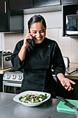 Positive young ethnic chef woman in uniform talking on cellphone while preparing delicious vegetable salad dish in kitchen of restaurant