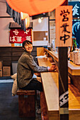 Asian woman in casual wear sitting at wooden counter while waiting for order in ramen bar