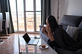 Konzentrierte Asiatin mit Heißgetränk in der Hand schaut auf den Bildschirm eines Netbooks auf dem Tisch mit Gebäck, während sie auf einer bequemen Couch sitzt
